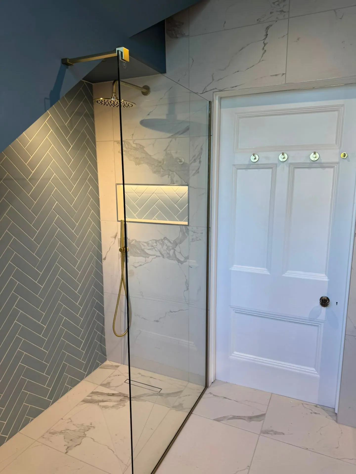 Walk-in shower with brass fittings, herringbone feature tiles and marble-effect walls by Heat Solution London