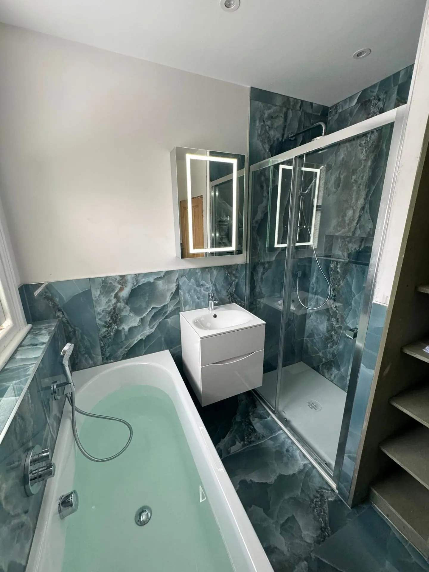 Dark marble-effect bathroom with bath, shower enclosure and illuminated mirror by Heat Solution London